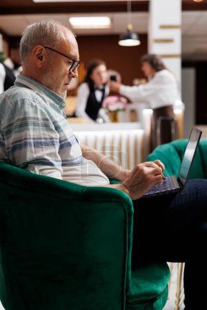 Photo for Caucasian senior male client relaxing on comfy sofa with personal computer in hotel lounge area. Retired senior man looking for enjoyable holiday activities on laptop in beautiful resort. - Royalty Free Image