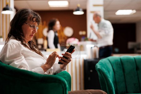 Photo for Retired senior woman enjoys online connectivity in hotel lobby, using mobile phone for communication and relaxation. Elderly female traveler relaxing with smartphone in luxury lounge area. - Royalty Free Image