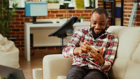 Téléchargez les photos : Passionné passionné homme afro-américain jouer à des jeux vidéo sur smartphone, se détendre après une longue journée au travail. Joueur compétitif enthousiaste s'amuser sur le téléphone dans le salon élégant appartement - en image libre de droit