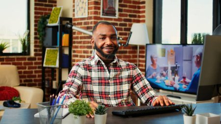 Téléchargez les photos : Homme afro-américain souriant optimiste profitant de son séjour à la maison travail indépendant, assis à un bureau confortable dans un élégant appartement chaleureux. Joyeux télétravailleur heureux travaillant à la maison, gros plan - en image libre de droit