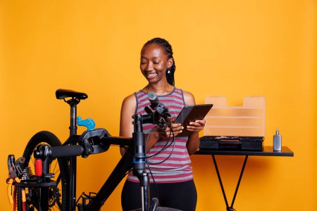 Téléchargez les photos : Femme afro-américaine active en bonne santé répare un vélo cassé avec sa boîte à outils, en utilisant une tablette intelligente pour la recherche et les instructions. Femme noire souriante utilisant un dispositif numérique pour l'entretien du vélo. - en image libre de droit