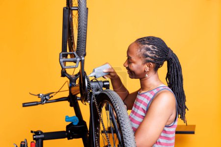 Téléchargez les photos : African American cycliste examine attentivement et répare les composants de vélo avec des outils spécialisés et lubrifiant pour une performance en douceur. Femme noire graissant et entretenant diligemment la chaîne de vélo. - en image libre de droit