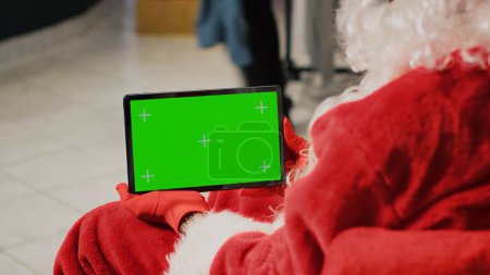 Téléchargez les photos : Employé dans le magasin de vêtements décorés de Noël habillé comme le Père Noël tenant la tablette d'écran vert, la mise en place d'articles de vêtements de site Web, la saisie des offres promotionnelles de Noël en ligne - en image libre de droit
