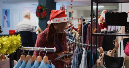 Photo for African american manager wearing Santa hat browsing through clothes racks at Christmas themed clothing store, using tablet to input reduced prices on online shop during holiday season sales - Royalty Free Image