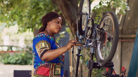 Téléchargez les photos : Portrait photo de femme noire enthousiaste inspectant soigneusement pour les dommages sur la roue de vélo pour l'entretien. Jeune femme afro-américaine réparer son propre vélo dans la cour, vérifier les pédales pour réparer. - en image libre de droit