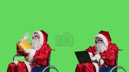 Téléchargez les photos : Saint Nick avec livre et ordinateur portable assis en fauteuil roulant, traitant de la déficience et la lecture de nouvelles ou la navigation sur la page Web. Jeune homme habillé en père Noël contre écran vert. - en image libre de droit