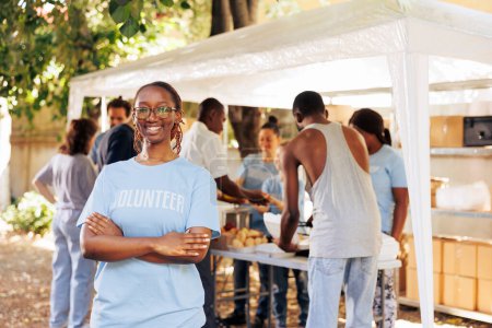 Junge schwarze Frau mit Brille steht draußen, die Arme verschränkt, und blickt in die Kamera. Vielfältige Gruppe von Freiwilligen unterstützt ein gemeinnütziges Programm, das sich der Hungerhilfe und der Hilfe für bedürftige Menschen widmet.