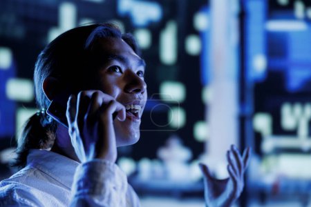 Photo for Happy man strolling around cityscape during nighttime, enjoying discussion over the phone with girlfriend, close up. Asian citizen making telephone calls while commuting, blurry background - Royalty Free Image