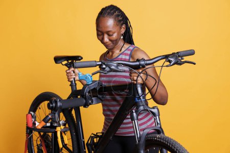 Foto de Ajuste y atlética mujer afroamericana hace reparaciones anuales de bicicletas con herramientas de trabajo. Mujer negra sana apoyando una bicicleta dañada en el soporte de reparación para el mantenimiento contra el fondo aislado. - Imagen libre de derechos