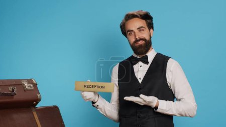 Photo for Hotel staff uses reception indicator on camera, guiding people in the right direction. Classy doorkeeper working in hospitality industry, presenting front desk pointer in studio to guide guests. - Royalty Free Image