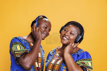 Foto de Sonriendo pareja romántica afroamericana usando ropa étnica disfrutando de la música juntos en auriculares inalámbricos. Hombre y mujer con expresión alegre escuchando canciones en estudio - Imagen libre de derechos