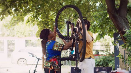 Photo for Young enthusiastic caucasian man and african american woman performing maintenance on bike wheels as outdoor summer hobby. Multiethnic couple fastening bicycle tire with professional equipments. - Royalty Free Image