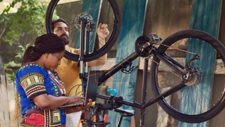 Photo for Vibrant african american lady browsing on internet for bicycle gear maintenance. Black woman checking for repair instructions on laptop while caucasian man dismantles broken bike wheel. - Royalty Free Image