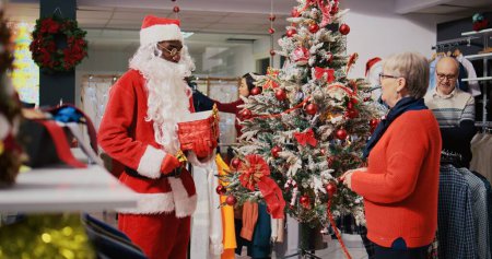 Foto de Empleado en Navidad adornan boutique de moda con traje de Papá Noel y los clientes se reunieron alrededor del árbol de Navidad bellamente decorado en la tienda de ropa durante la temporada de vacaciones de invierno - Imagen libre de derechos