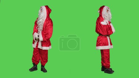 Téléchargez les photos : Santa Claude cosplay vérifie montre-bracelet, en regardant le temps et être impatient sur fond d'écran vert du corps entier. Père Noël se promenant à la caméra, célébration saisonnière. - en image libre de droit