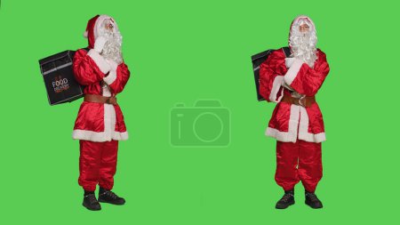 Foto de Santa Claus en traje rojo con mochila trabajando como repartidor durante la víspera de Navidad, fondo de pantalla verde de cuerpo completo. Saint Nick Cosplay entrega de pedidos de comida rápida para las vacaciones de invierno. - Imagen libre de derechos