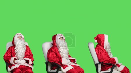 Photo for Santa claus character on chair against greenscreen backdrop, cheerful person celebrating christmas eve in red costume. Young adult advertising december holiday with iconic winter suit. - Royalty Free Image