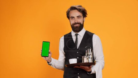 Photo for Young waiter shows greenscreen on camera, holding smartphone with blank copyspace display. Friendly elegant fine dining employee indicates isolated empty screen template in studio. - Royalty Free Image