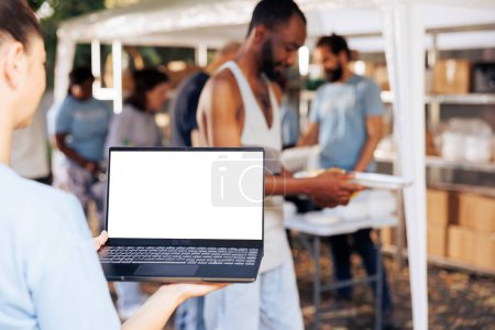 Photo for Caucasian woman working for charity is shown in a close-up holding a laptop with a blank mockup template. Female volunteer grasping a minicomputer with a customizable copyspace white screen. - Royalty Free Image