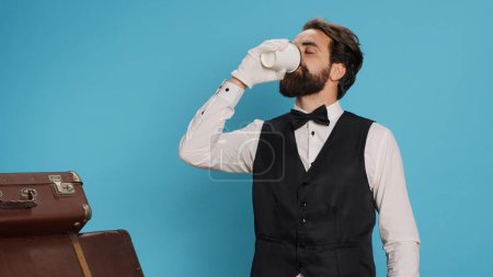 Photo for Hotel porter drinking coffee on camera, bellboy wearing luxurious clothes and white gloves. Doorman employee enjoying drink next to suitcases and trolley bags against blue background. - Royalty Free Image