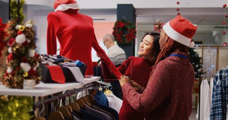 Amable asistente de venta al por menor en el centro comercial adornado festivo tienda de moda que muestra al cliente hermosas prendas rojas, listas para ser usadas en eventos navideños temáticos de Navidad durante la temporada de invierno.