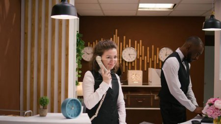 Photo for Hotel staff answers calls at reception, using landline phone to make remote bookings at front desk. Friendly receptionist keeping evidence of accommodations and room reservations. - Royalty Free Image