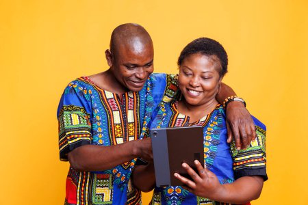 Photo for Smiling wife and husband with joyful expression hugging while using digital tablet together. Cheerful woman holding portable gadget while man tapping on touchscreen in studio - Royalty Free Image