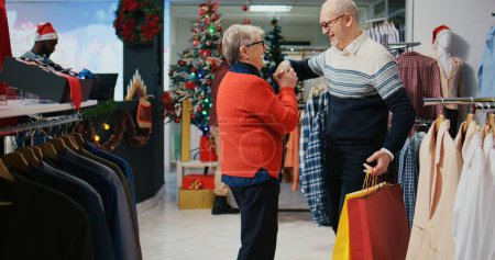Foto de Hombre mayor sosteniendo bolsas de compras, esposa girando en la tienda de moda decorada festiva, feliz después de encontrar regalos ideales para compartir con los miembros de la familia en la fiesta de Navidad - Imagen libre de derechos