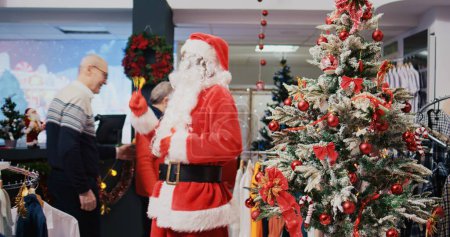 Foto de Alegre trabajador afroamericano vestido como Santa Claus en la tienda de ropa adornada de Navidad centro comercial. Empleado jingling campanas de Navidad y saludar a los clientes felices en la tienda de moda adorno festivo - Imagen libre de derechos