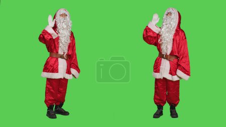 Photo for Friendly character wave and greet people on camera, wearing traditional festive costume to spread christmas spirit. Young adult dressed as santa claus saying hello, greenscreen backdrop. - Royalty Free Image