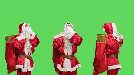 Téléchargez les photos : Nick saint fatigué bâillant sur fond vert, se sentant épuisé de porter sac avec des cadeaux et des jouets pour les vacances de la veille de Noël. Mode de réalisation du Père Noël somnolent en studio, costume rouge. - en image libre de droit