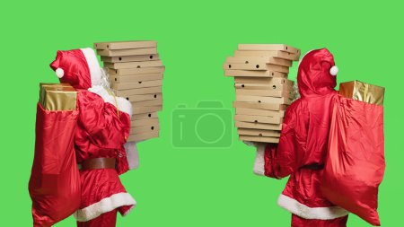 Photo for Character delivering boxes of pizza in studio, standing over greenscreen with big pile of fast food and bag filled with presents. Santa claus in seasonal winter costume acting like a deliveryman. - Royalty Free Image