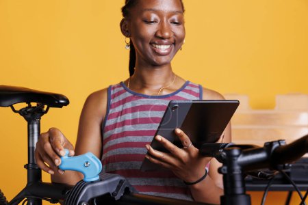 Téléchargez les photos : Vélo de fixation féminin afro-américain sportif avec outils et dispositif numérique de navigation pour des solutions. Plan détaillé de femme noire en bonne santé à la recherche sur tablette téléphonique pour l'entretien du vélo. - en image libre de droit