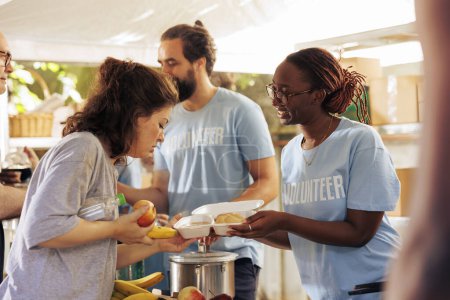Foto de Mujer voluntaria afroamericana en el centro local que participa activamente en la distribución de comidas nutritivas para aliviar el hambre entre los menos afortunados. Mujer negra ayudante compartiendo comida con personas sin hogar. - Imagen libre de derechos