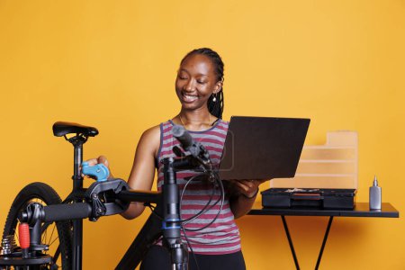 Photo for African American female inspecting and repairing bike with specialized tools and digital instructions from wireless computer. Active black woman examining and servicing broken bicycle with laptop. - Royalty Free Image
