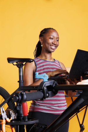 Téléchargez les photos : Focalisé jeune femme répare vélo cassé, en utilisant des outils spécialisés et la recherche de solutions d'entretien sur ordinateur portable. Vibrante navigation féminine afro-américaine sur Internet pour les nouvelles pièces de vélo modernes. - en image libre de droit
