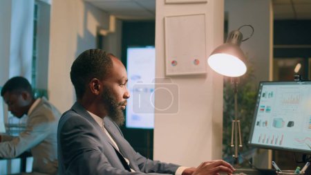 Téléchargez les photos : Des hommes d'affaires afro-américains assis au bureau, regardant les tableaux financiers, imputant des données sur ordinateur pendant le quart de nuit. Coworkers résoudre les tâches du projet de l'entreprise nuit avant l'échéance - en image libre de droit