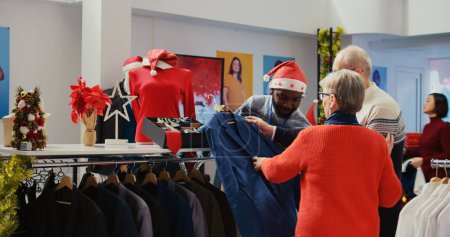 Photo for Wife and husband in xmas ornate shopping mall fashion shop, being assisted by african american worker with finding ideal outfit during festive holiday season promotional sales - Royalty Free Image