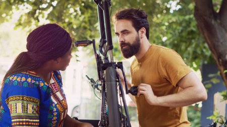 Photo for Dedicated multiracial pair cautiously fastening bike chain and pedal for summer outdoor leisure cycling. Outdoorsy young couple thoroughly examining damages on bicycle wheel for repairing. - Royalty Free Image