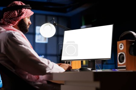 Téléchargez les photos : L'image représente un homme musulman assis à son poste de travail, son écran d'ordinateur affichant un gabarit chromatique isolé. Jeune homme arabe travaillant sur un ordinateur de bureau avec un écran blanc copyspace. - en image libre de droit