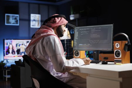 Photo for Dedicated software programmer wearing traditional Arabic clothing is seated at his home office desk examining database code on computer. Muslim man checking the programming language on the pc monitor. - Royalty Free Image