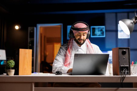 Téléchargez les photos : Homme arabe confiant tapant sur un ordinateur portable engagé dans la communication, la vidéoconférence et le courrier électronique. Musulman écoute et regarde avec casque sans fil tout en recherchant et en naviguant sur Internet. - en image libre de droit