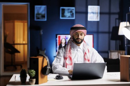 Foto de Un hombre de negocios árabe de confianza trabajando en un escritorio moderno, escribiendo en su portátil. Chico musulmán dedicado a la navegación, la comunicación por correo electrónico y la conectividad inalámbrica en un entorno de oficina tranquilo y profesional. - Imagen libre de derechos