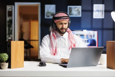 Photo for A traditional Middle Eastern entrepreneur diligently works in a modern office, utilizing wireless technology for communication and research. Young Arab man using his wireless headphones and laptop. - Royalty Free Image