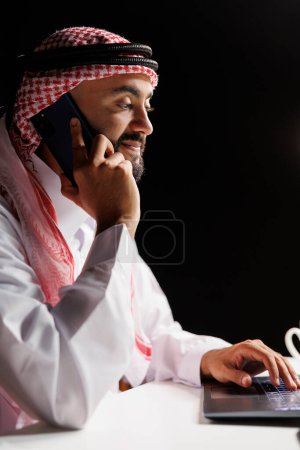Foto de Hombre de Oriente Medio, vestido tradicionalmente, comprometido en una conversación telefónica, que multitareas con su dispositivo móvil y portátil. Una mezcla moderna de patrimonio y tecnología. Vista lateral, primer plano. - Imagen libre de derechos