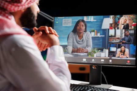 Foto de La imagen muestra la pantalla de la computadora mostrando una reunión en línea entre personas multiétnicas. Vista detallada de un hombre de negocios musulmán con atuendo tradicional participando en una videollamada. - Imagen libre de derechos