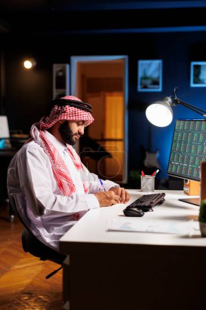 Téléchargez les photos : Arabe gars absorbé dans son travail, la recherche, la prise de notes, et surfer sur le net. En utilisant son ordinateur portable et son moniteur, l'homme musulman professionnel fait preuve d'engagement et de professionnalisme. - en image libre de droit