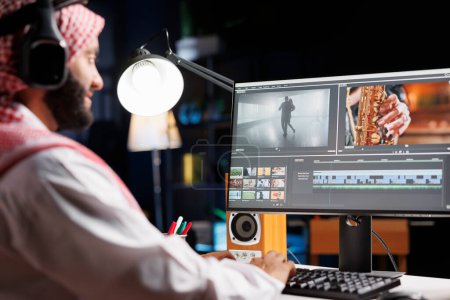 Photo for Arab male filmmaker seated at table diligently working on post production for movie. Muslim video editor in traditional attire, wearing wireless headphones while examining footage on computer screen. - Royalty Free Image