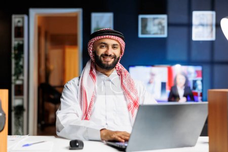 Foto de Joven vestido con ropa árabe usando su portátil en la mesa. El musulmán, confiado y conocedor de la tecnología, sonríe a la cámara, simbolizando la mentalidad empresarial de la era digital. - Imagen libre de derechos