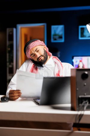 Photo for Detailed shot of Arab man using a laptop and mobile device for communication and research, showcasing proficiency in technology. Muslim guy multitasking, talking on his cellphone and comparing notes. - Royalty Free Image